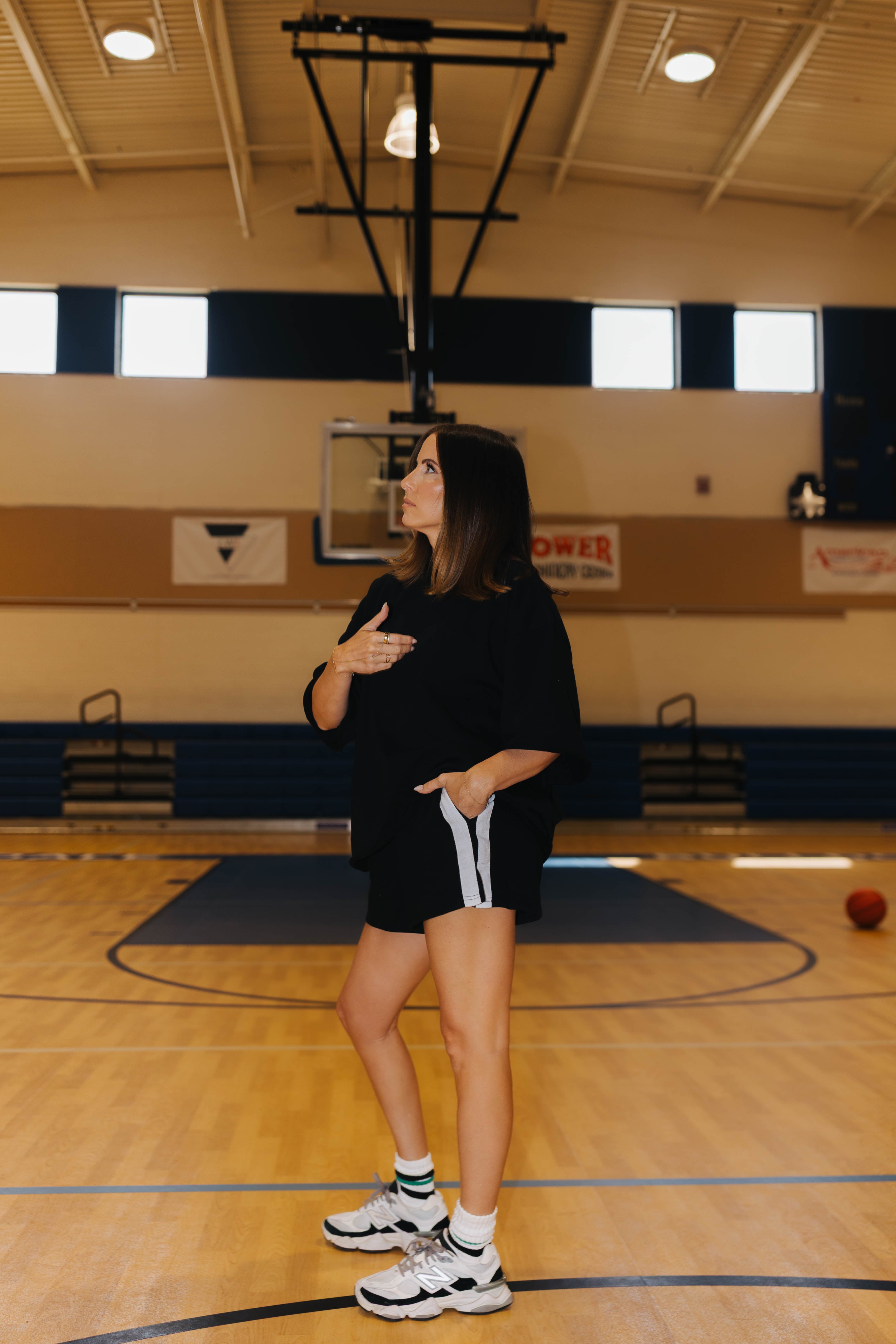 A woman stands on a basketball court wearing a black oversized t-shirt and black athletic shorts with white side stripes, paired with white sneakers.