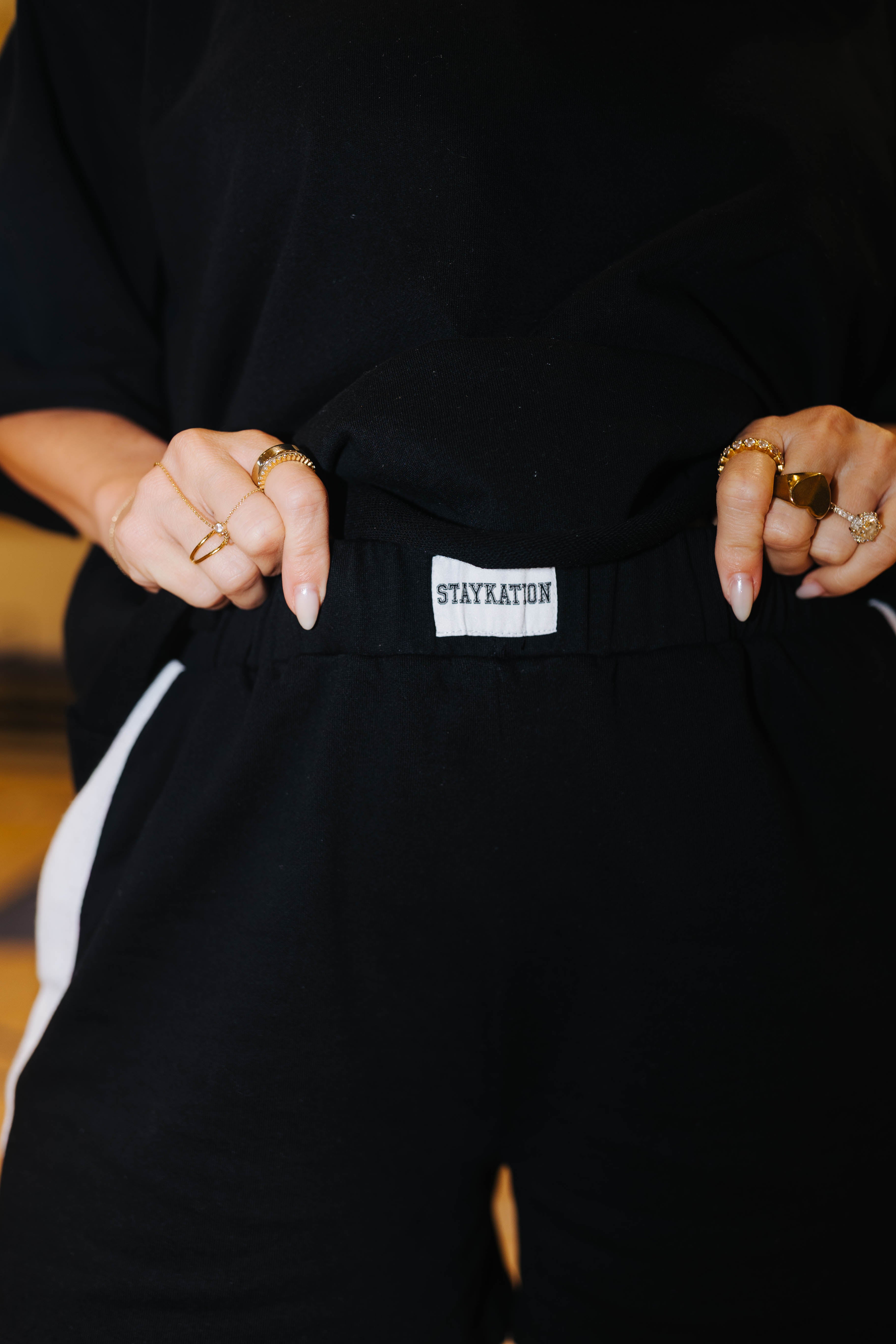 Close-up of a person wearing black shorts with a white side stripe and a visible white brand tab reading STAYKATION at the waistband, paired with a black top.