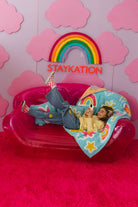Person lying on a pink inflatable chair with a rainbow and 'STAYKATION' text against a pink cloud-patterned background.