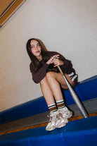 A young woman sits on a blue gym bleacher, holding a metal baseball bat. She is wearing a dark brown long-sleeve sweatshirt set 
