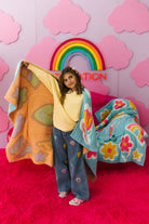Person holding colorful blankets against a pink wall with a rainbow and clouds.