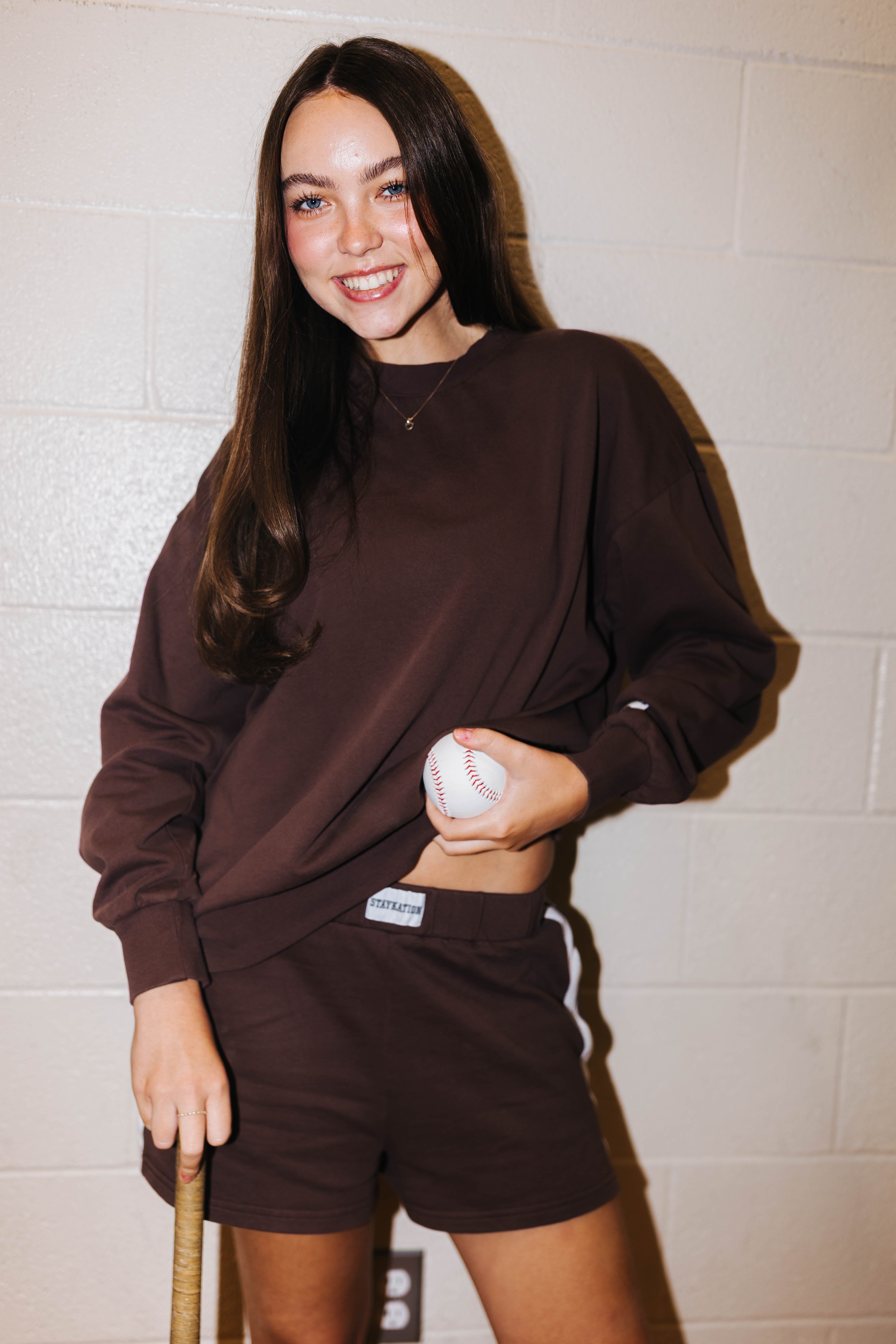 A smiling young woman stands against a light cinderblock wall wearing a matching dark brown sweatshirt and shorts set, holding a baseball in one hand and a bat in the other.