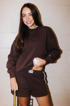 A smiling young woman stands against a light cinderblock wall wearing a matching dark brown sweatshirt and shorts set, holding a baseball in one hand and a bat in the other.