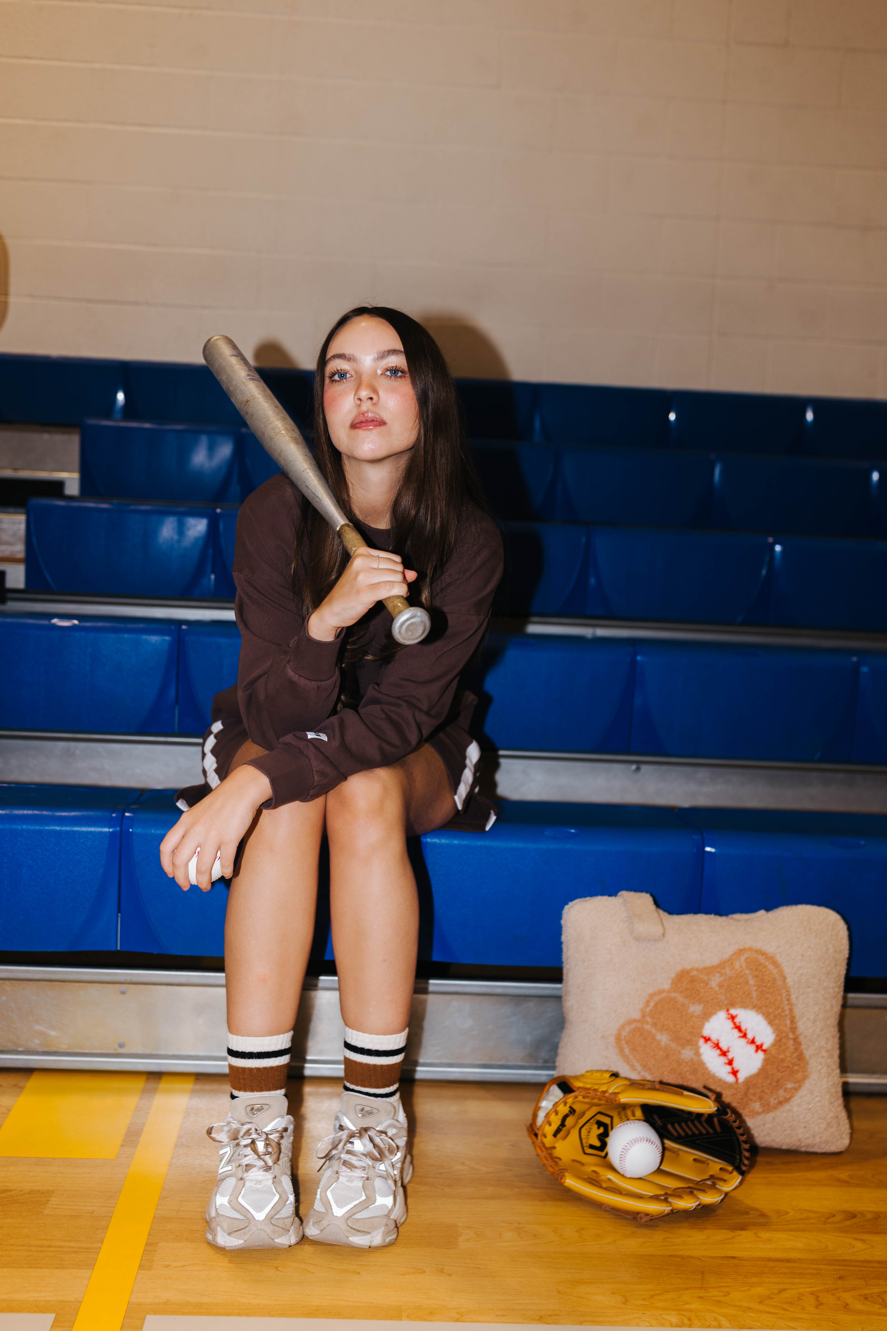 A young woman sits on blue gym bleachers holding a baseball bat, wearing a dark brown sweatshirt, shorts, and white sneakers. 