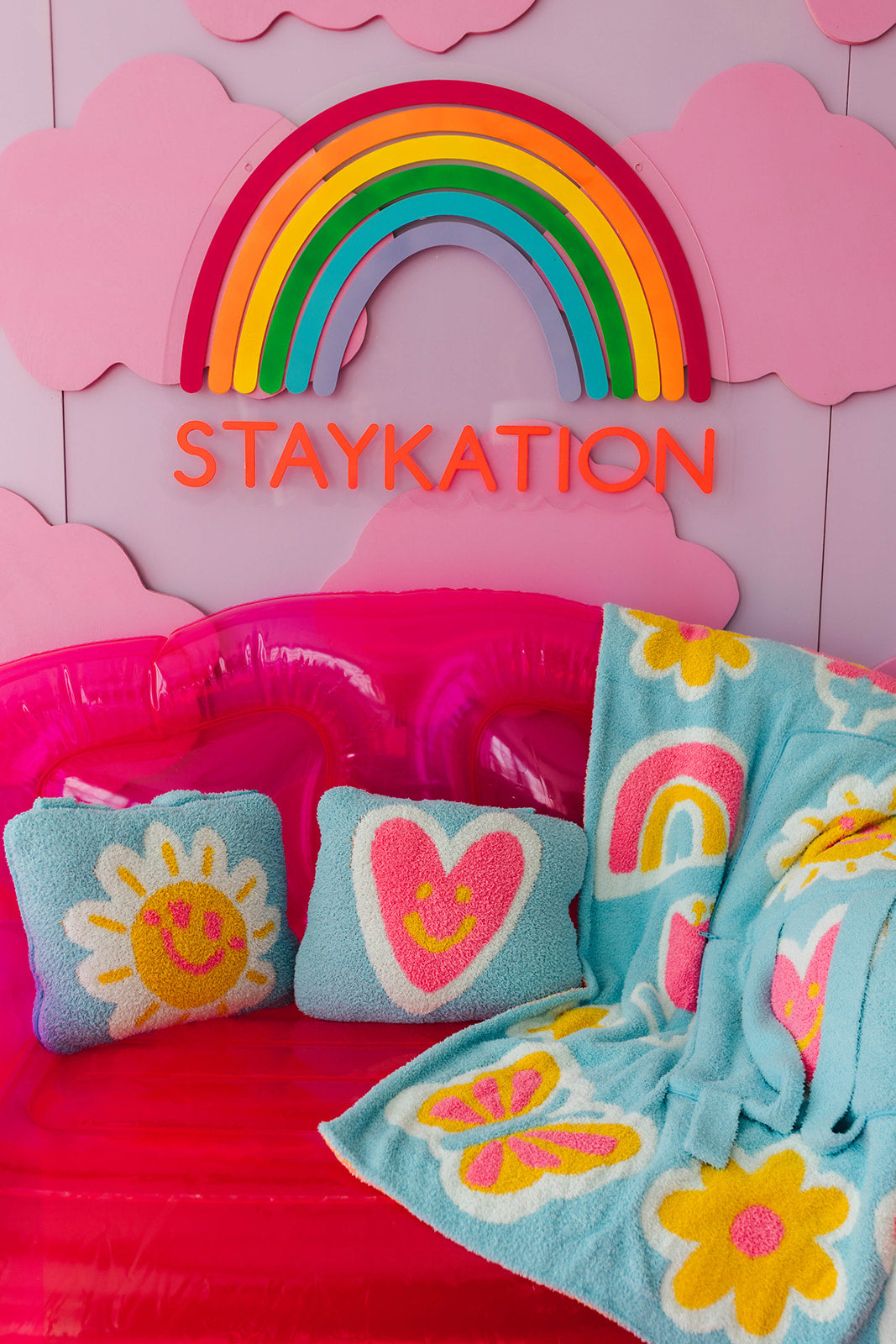 Children's inflatable bed with colorful pillows and blanket, featuring a rainbow and 'STAYKATION' text on a pink background.