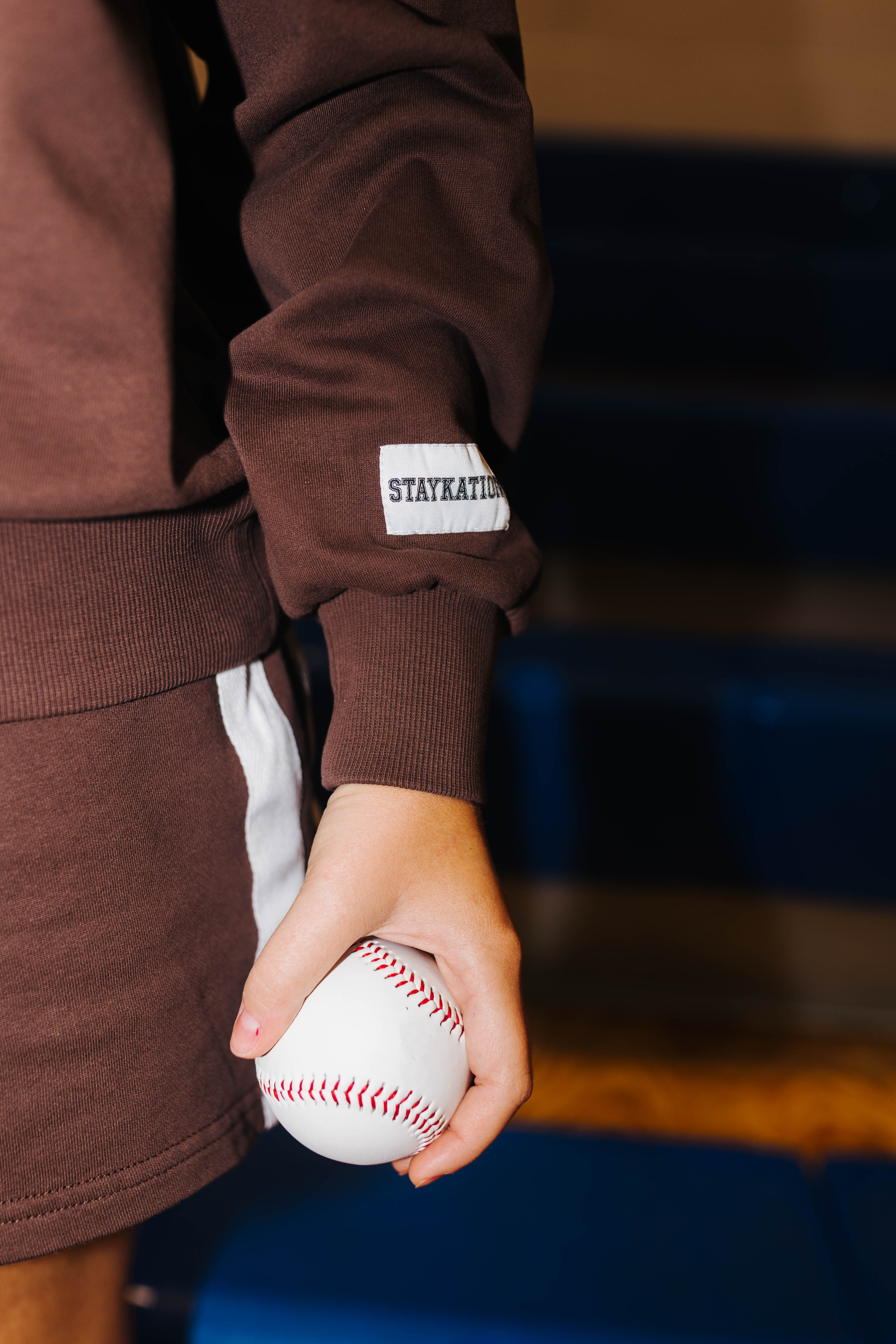 A close-up of a person holding a baseball, wearing a brown sweatshirt and shorts set with a white sleeve patch that reads 'STAYKATION'.