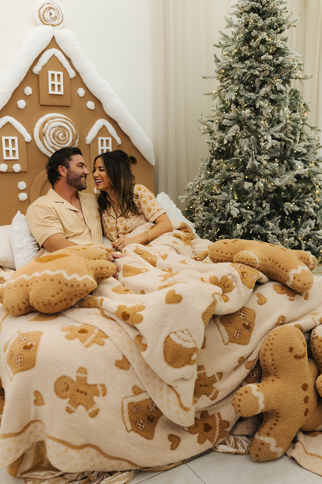 Couple under a gingerbread-themed blanket in front of a Christmas tree and gingerbread house.