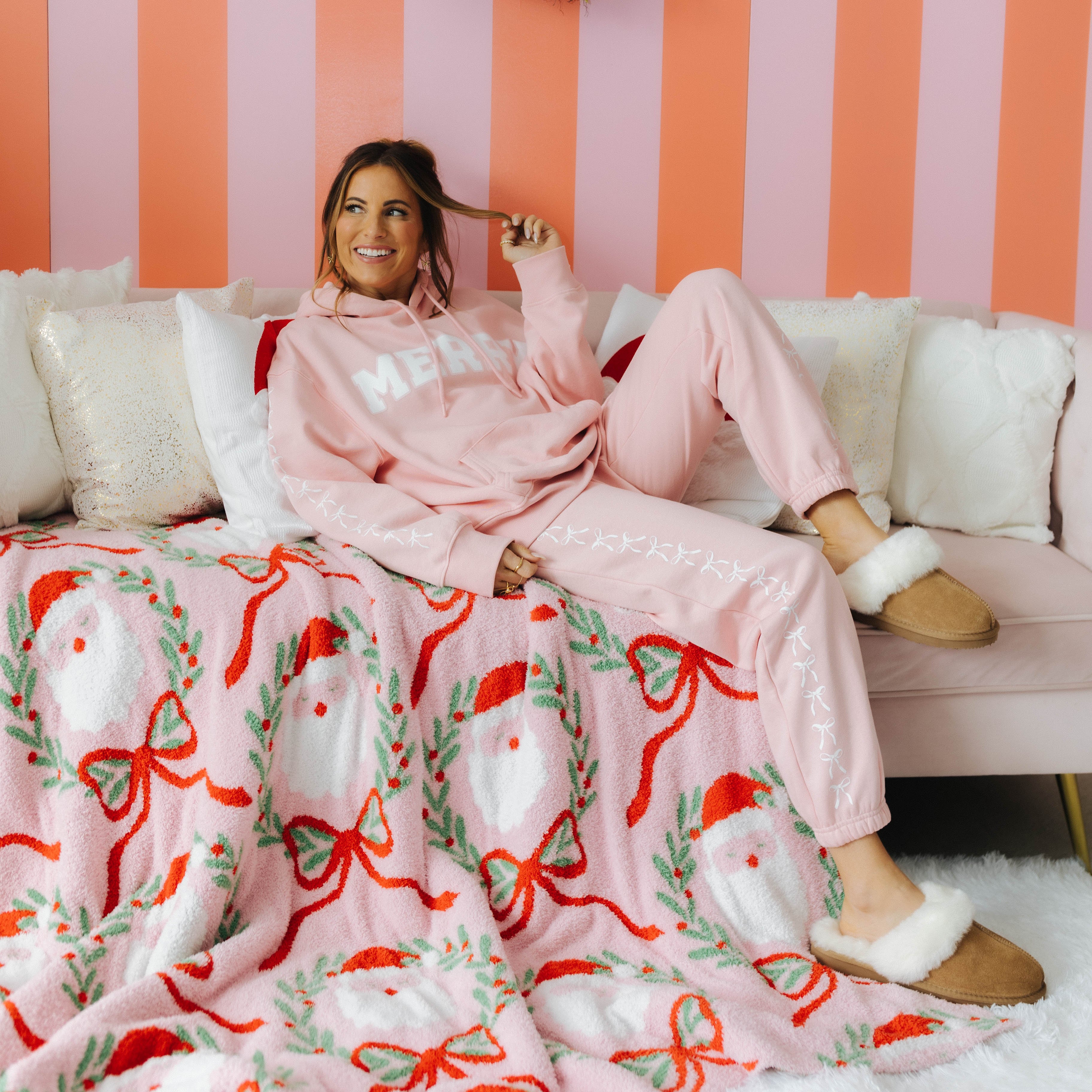 Woman in pink pajamas sitting on a couch with a Christmas-themed blanket and wreaths on a striped wall.