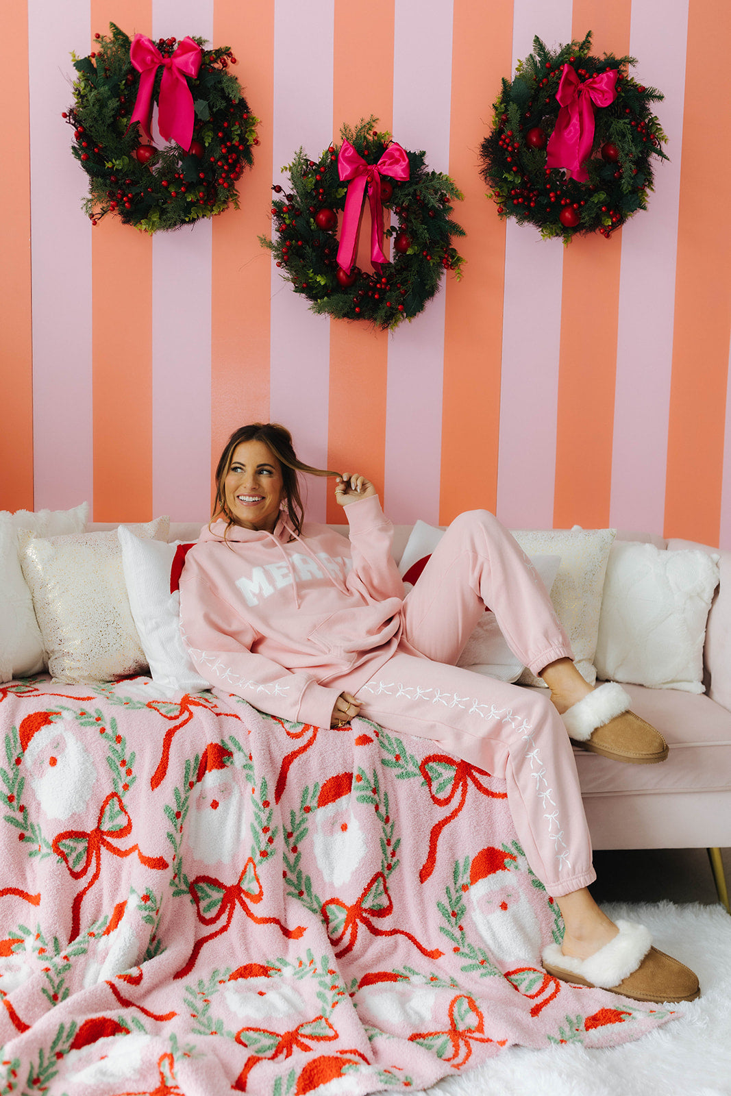 Woman in pink pajamas sitting on a couch with a Christmas-themed blanket and wreaths on a striped wall.