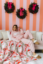 Woman in pink pajamas sitting on a couch with a Christmas-themed blanket and wreaths on a striped wall.