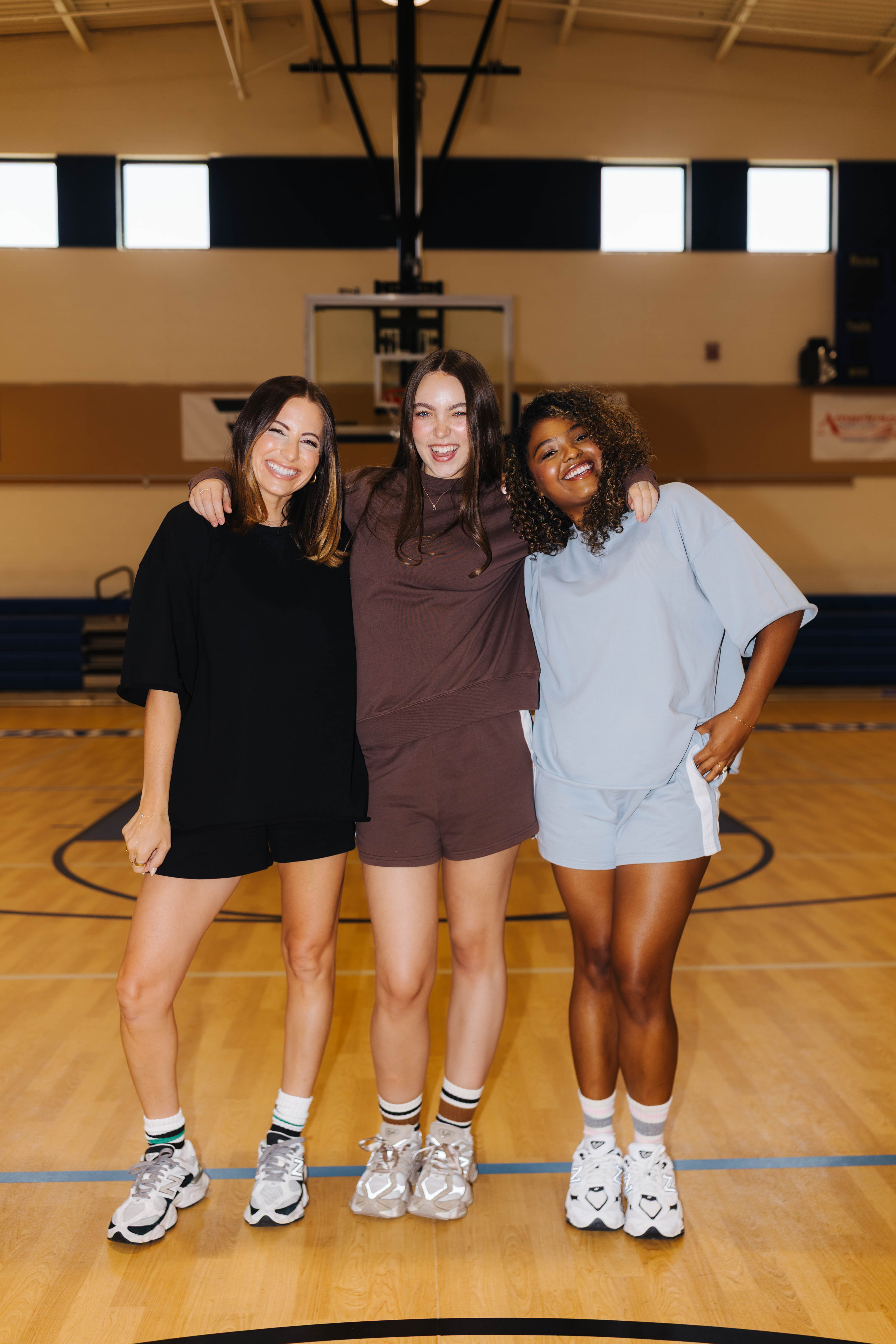 Three women posing together on a wooden basketball court, each wearing a matching oversized lounge set in black, brown, and light blue. The background shows a gymnasium with a basketball hoop and court markings.