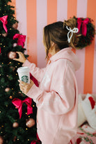 Woman in a pink hoodie holding a Starbucks cup, standing next to a Christmas tree with pink bows.