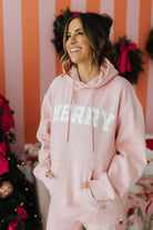 Woman wearing a pink 'Merry' hoodie in a festive setting with Christmas decorations.