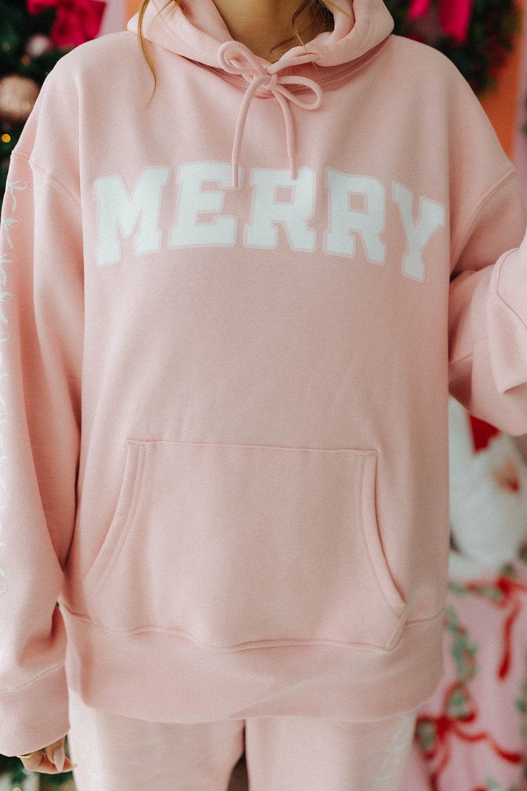 Pink hoodie with 'MERRY' text worn by a person, Christmas tree and decorations in the background