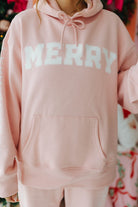 Pink hoodie with 'MERRY' text worn by a person, Christmas tree and decorations in the background
