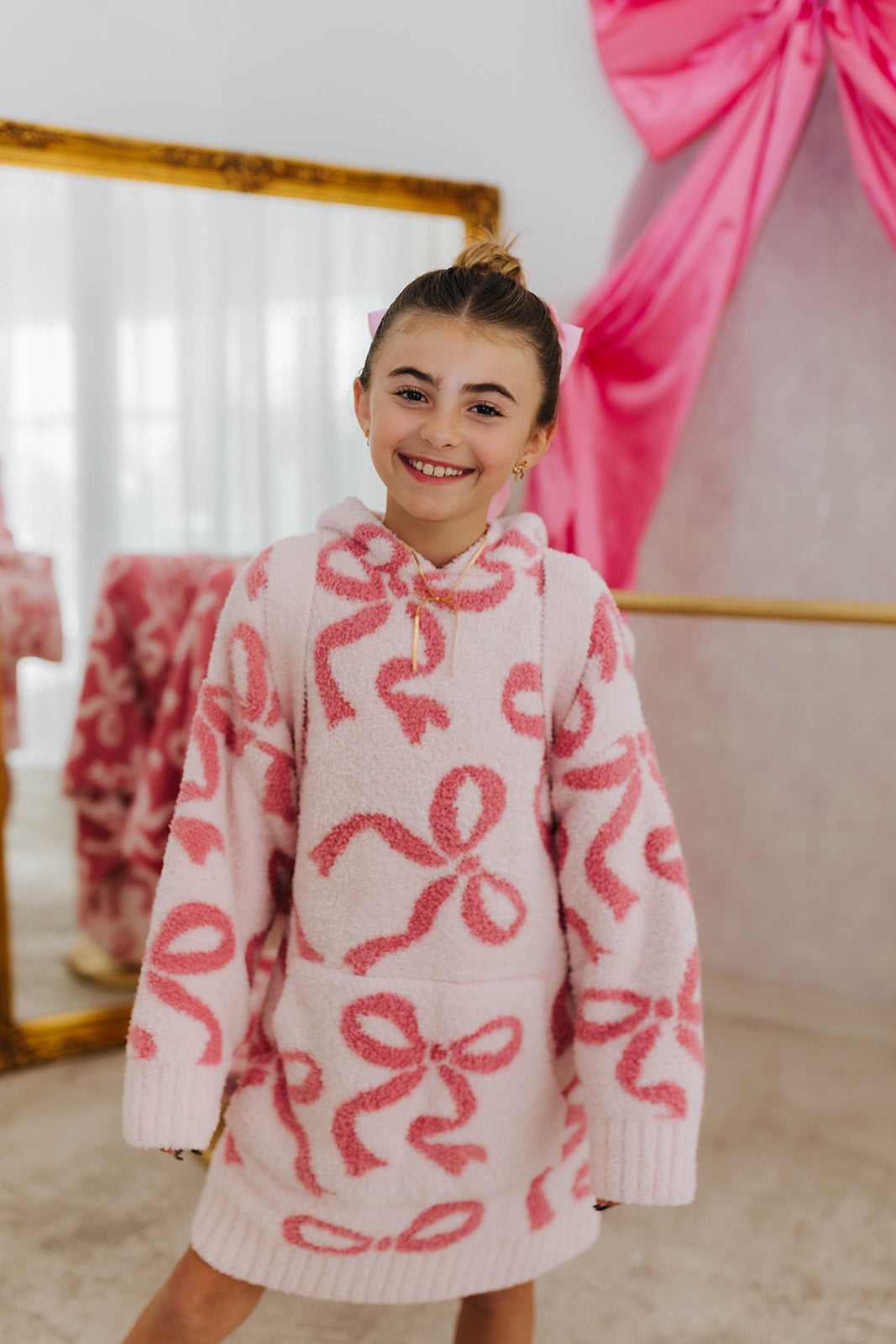 Person wearing a pink sweater with bow patterns in a room with a mirror and pink fabric.