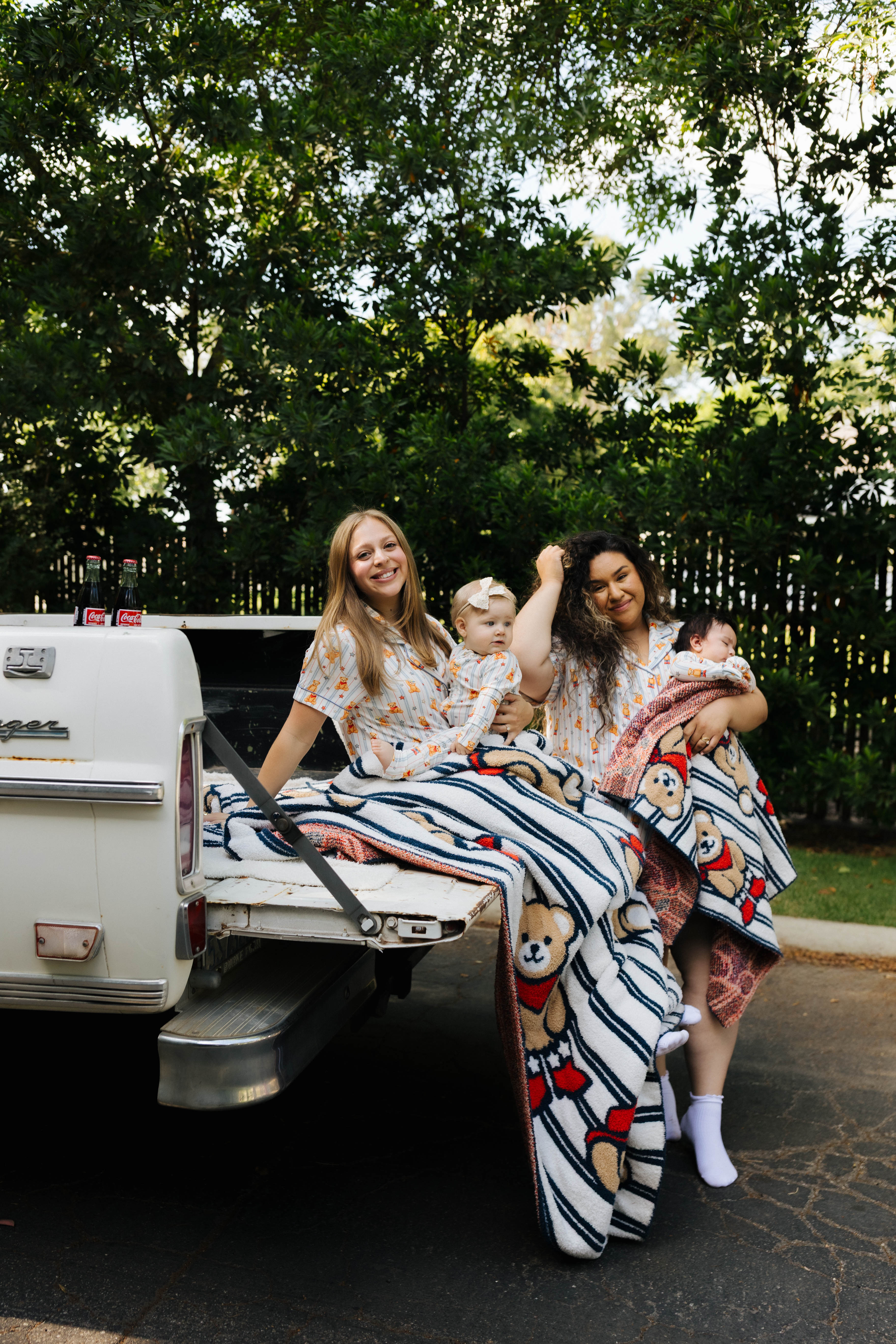 Two women with children sitting in the back of a white truck surrounded by greenery.