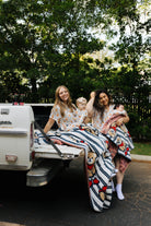 Two women with children sitting in the back of a white truck surrounded by greenery.