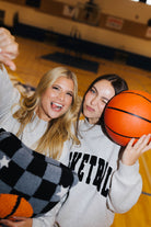 Two women posing together on a basketball court, both wearing light gray sweatshirts with black lettering. One holds an orange basketball, and the other holds a black, gray, and white checkered quillow.