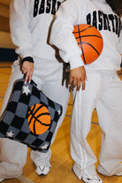 Two people in matching sweatpants and sweatshirts standing on a wooden gym floor. One holds a black-and-gray checkered quillow with an orange basketball graphic, and the other holds an orange basketball.