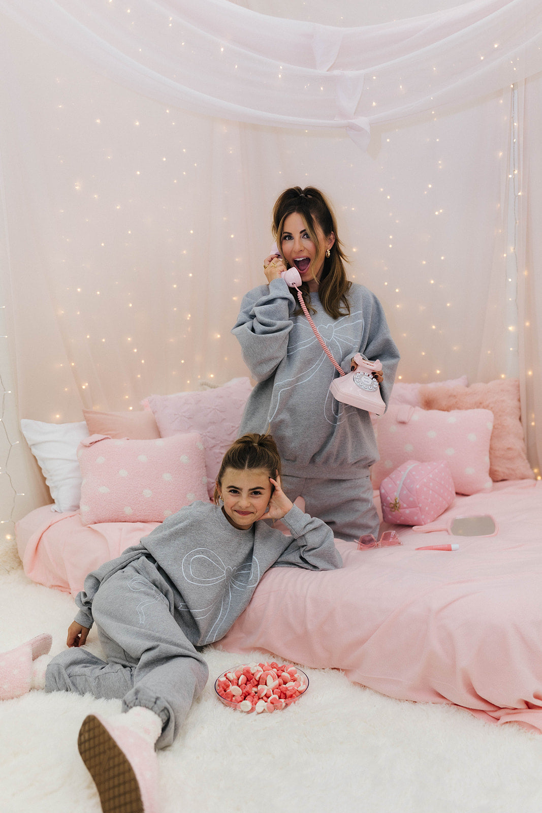Two people in matching grey outfits on a pink bed with fairy lights in the background