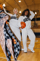 Two women posing on a basketball court wearing matching white sweatpants and sweatshirts with 'BASKETBALL' printed in black. One holds an orange basketball, and a patterned blanket is draped in the foreground.