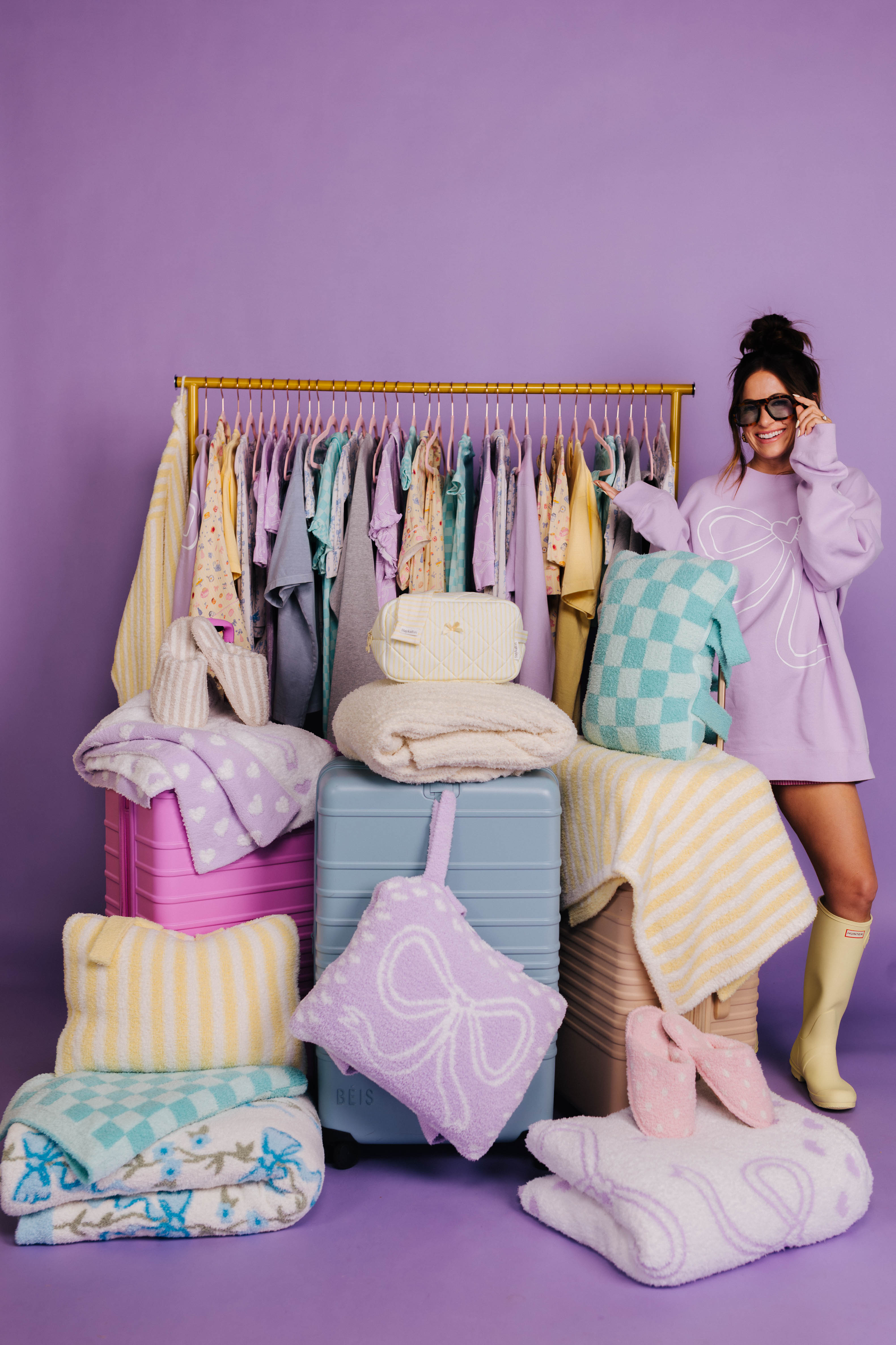 Woman in a pastel outfit standing next to a clothing rack with colorful clothes and accessories on a purple background