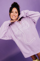 Woman wearing a light purple sweatshirt with a large white bow design on a purple background