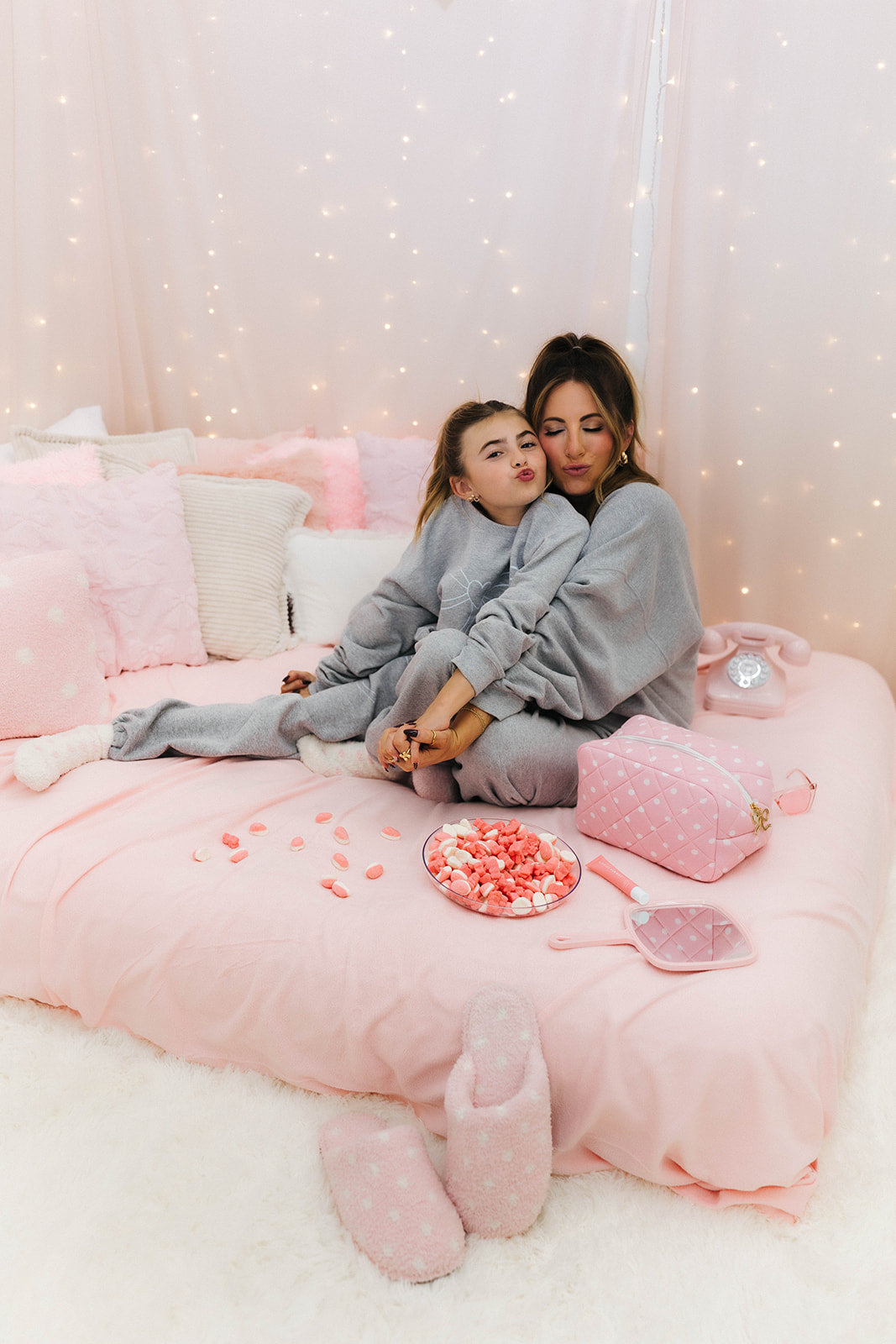 Two people in matching gray outfits sitting on a pink bed with decorative pillows and fairy lights.