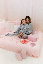 Two people in matching gray outfits sitting on a pink bed with decorative pillows and fairy lights.