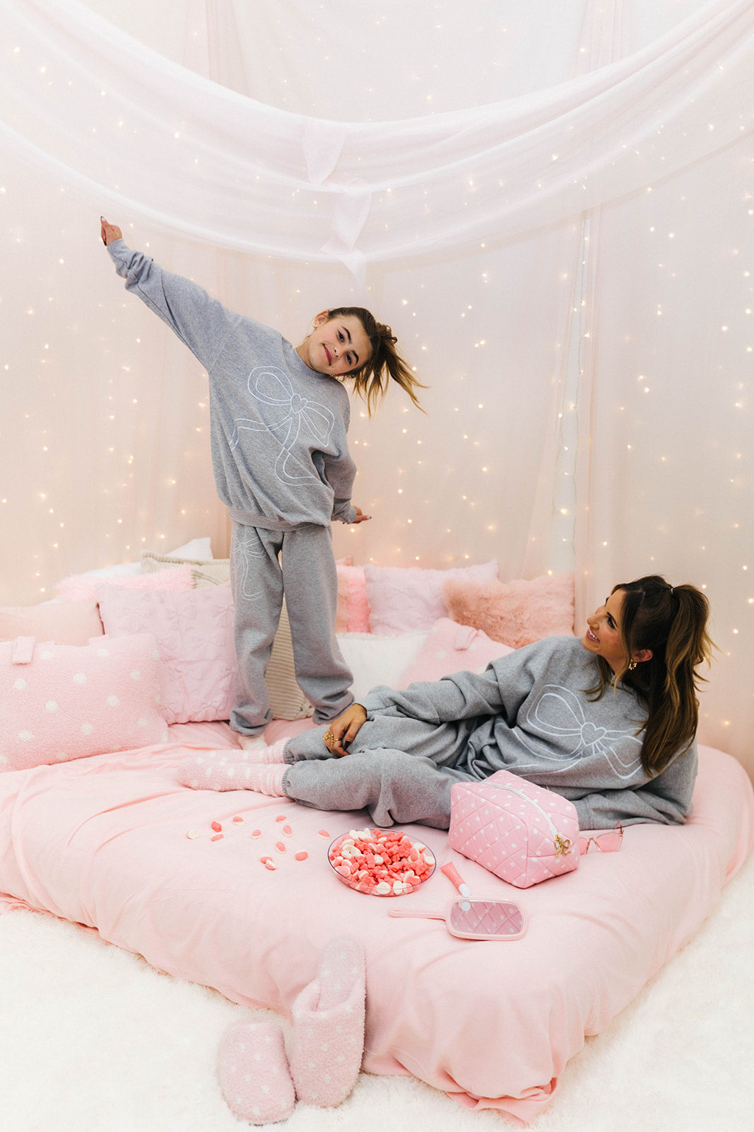 Two people in matching gray outfits on a pink bed with decorative pillows and fairy lights.