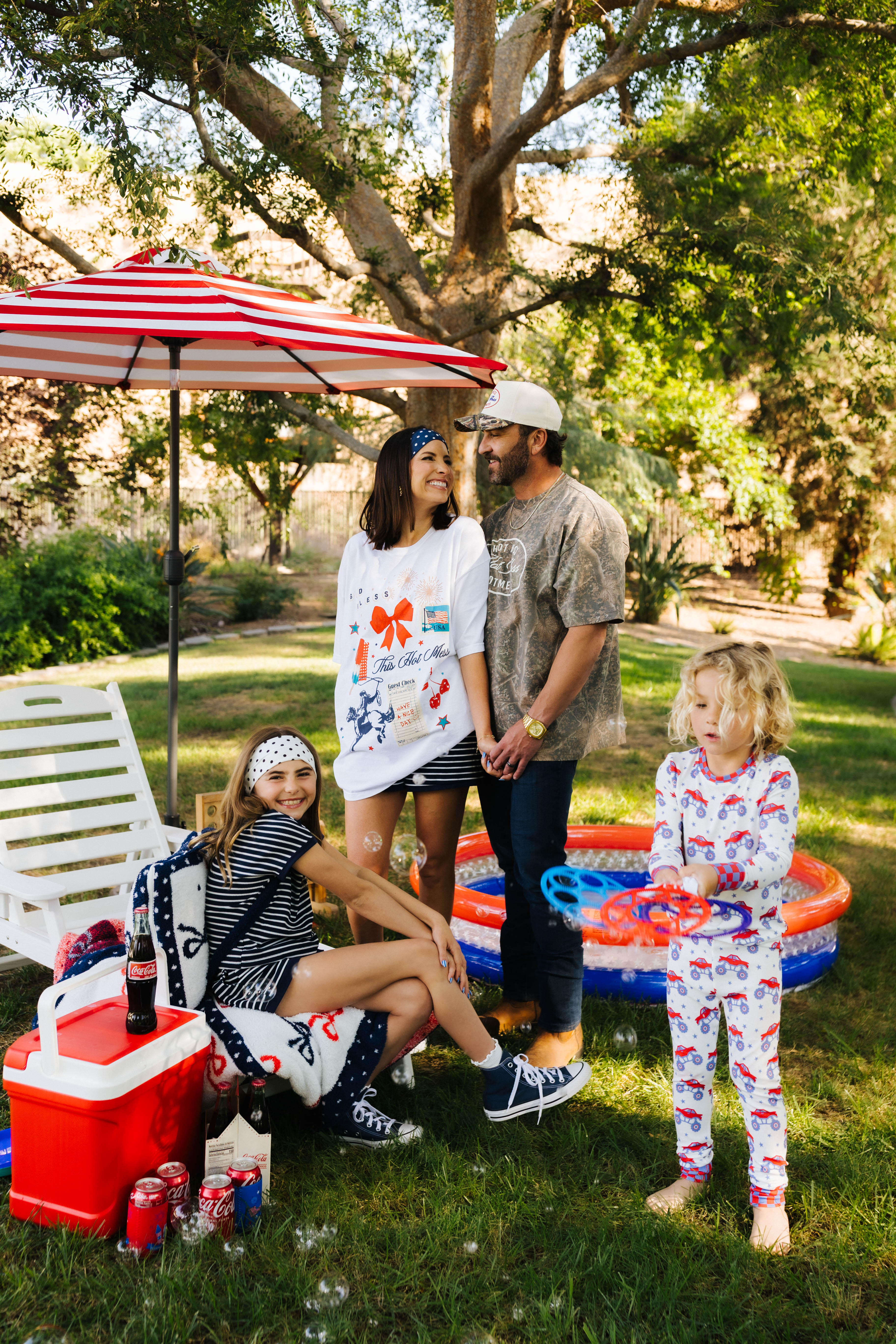 Family of four enjoying a sunny day outdoors with a red, white, and blue striped umbrella and pool.