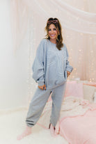 Woman wearing a grey sweatshirt and pants set in a cozy room with pink bedding and string lights.