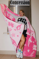 A woman wearing a gray 'CHEER' sweatshirt and black shorts stands indoors while holding up a large pink and white blanket with cheer silhouettes and stars. A sign reading 'Concession' is visible behind her.