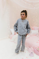 Child wearing a gray outfit with a bow design in a cozy room with pink bedding and fairy lights.