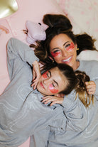 Two people wearing matching gray sweatshirts with heart designs on their faces, lying on a pink surface.