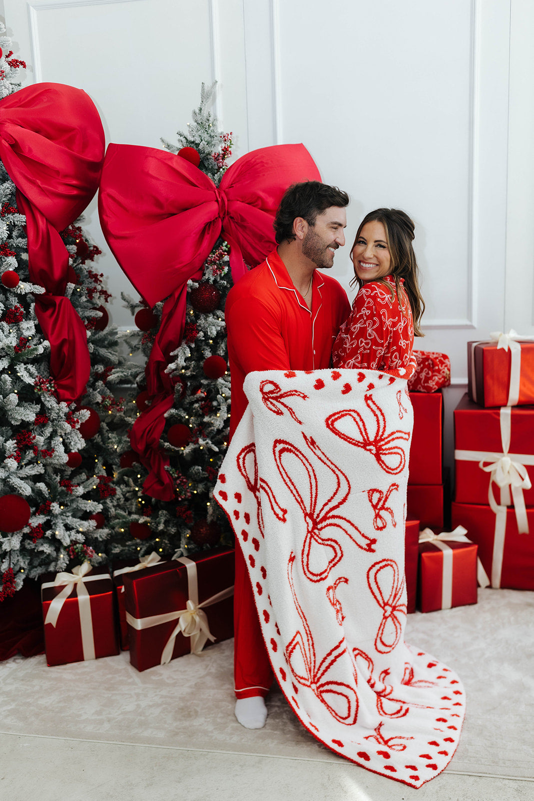 Couple in red pajamas standing in front of a Christmas tree with presents and a decorative blanket.
