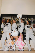 Four women in matching light gray football sweatpants and sweatshirts with 'FOOTBALL' printed in black, posing together in a room with sports trophies in the background. They are holding footballs and cheer pom-poms, with two of them carrying large decorative pillows.