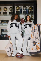 two girls in front of a trophee wall wearing matching football sweatshirts, holding a football blanket and football quillow