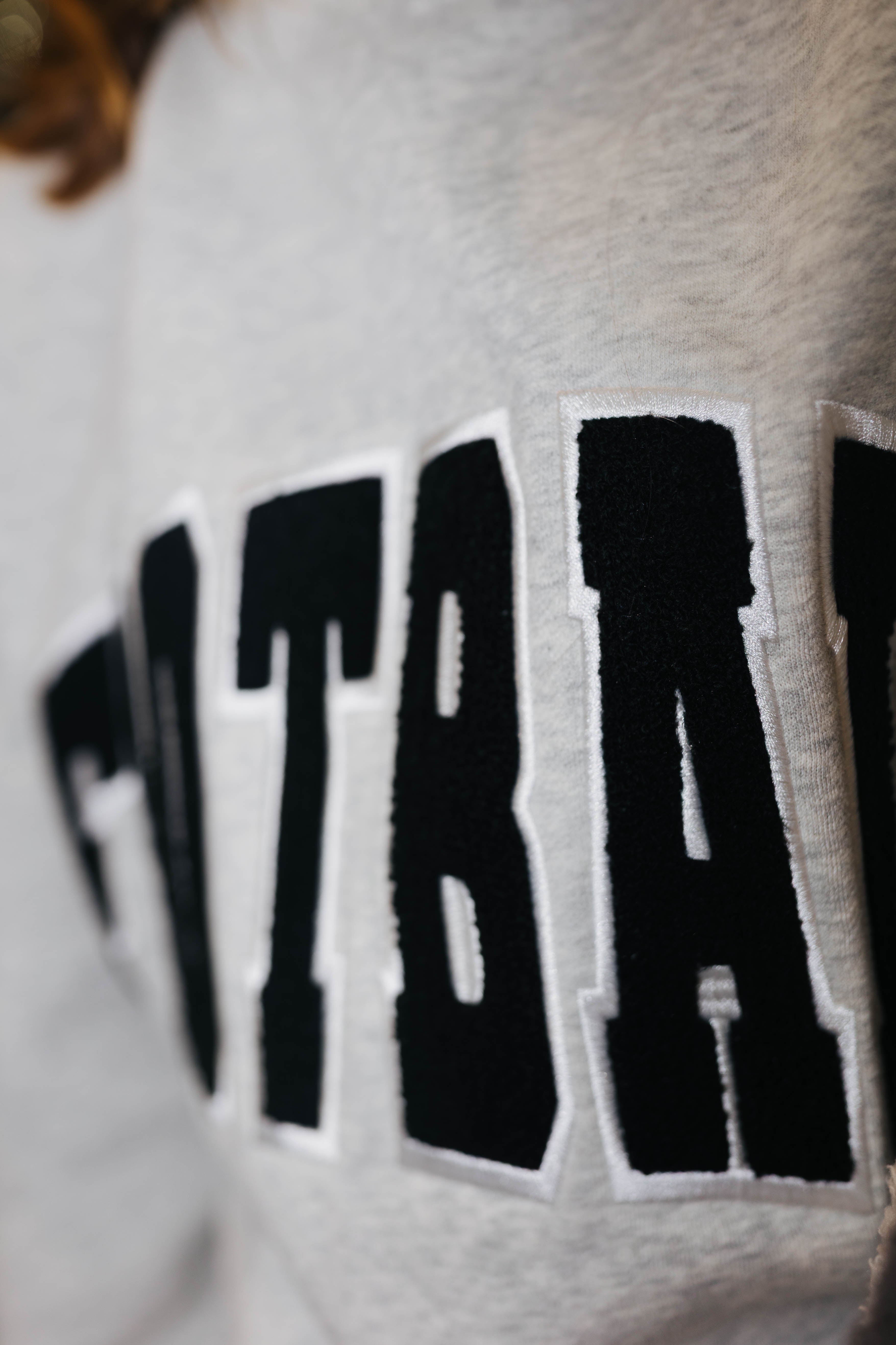 Close-up of a light gray heather sweatshirt featuring a large black collegiate-style embroidered patch with white border and the letters.