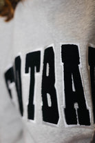 Close-up of a light gray heather sweatshirt featuring a large black collegiate-style embroidered patch with white border and the letters.