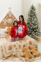 Two people wearing red Christmas sweaters with gingerbread-themed designs, sitting on a bed with gingerbread decorations and a Christmas tree in the background.