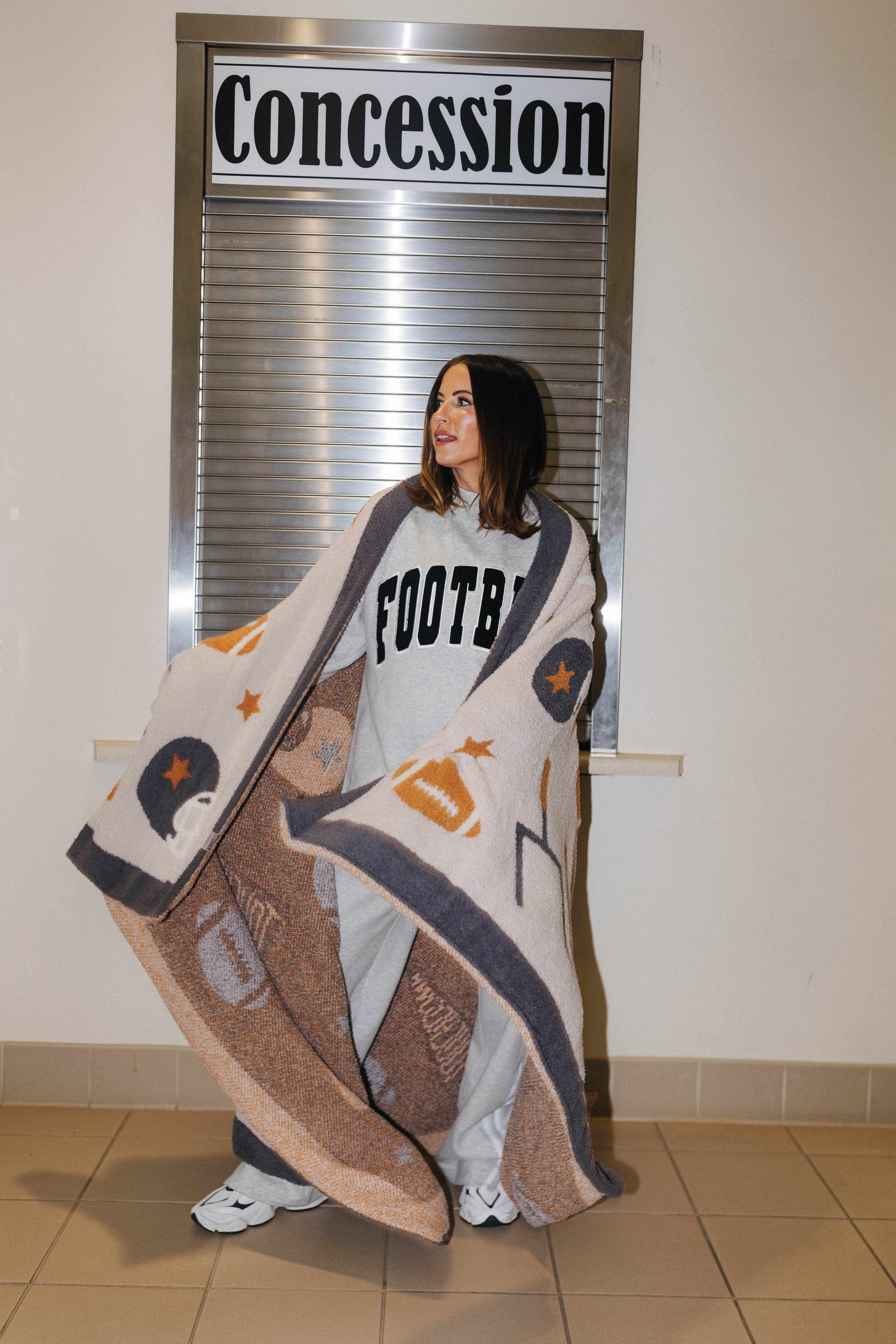 A woman stands indoors in front of a window with a 'Concession' sign, wrapped in a large beige and brown football-themed blanket with grey and orange accents. She is wearing a light gray sweatshirt with 'FOOTBALL' printed on it.