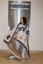 A woman stands indoors in front of a window with a 'Concession' sign, wrapped in a large beige and brown football-themed blanket with grey and orange accents. She is wearing a light gray sweatshirt with 'FOOTBALL' printed on it.