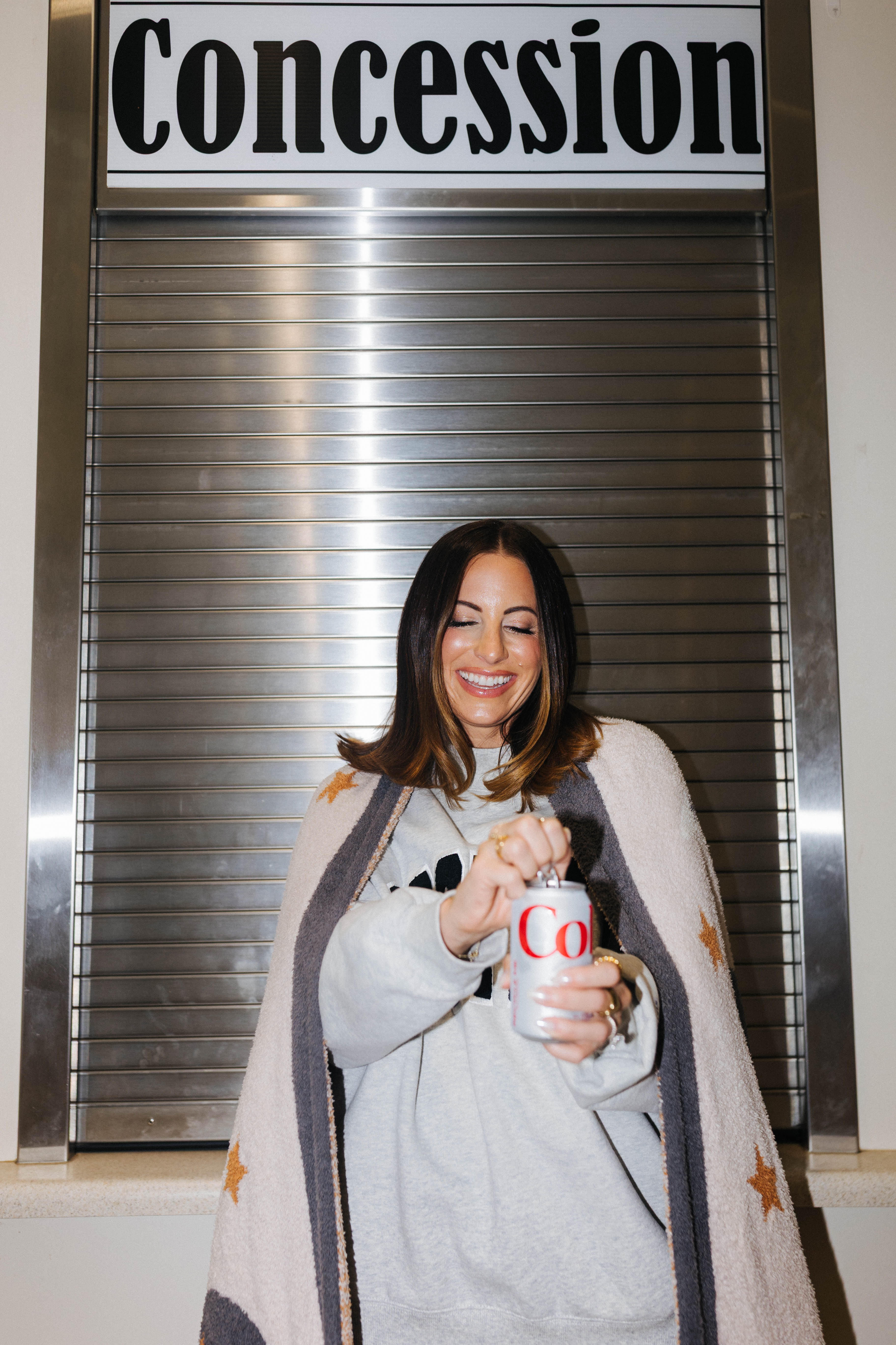 A smiling woman wrapped in a blanket with gold stars and dark trim stands in front of a closed metal roll-up door under a sign that reads 'Concession.' She holds a white can with red lettering.