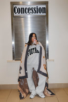 A woman stands in a hallway wrapped in a large beige and brown patterned blanket with 'FOOTBALL' printed on a light gray sweatshirt draped over her shoulders. A 'Concession' sign is behind her.