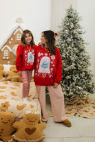 Two people wearing matching red sweaters with Christmas designs in a festive room with a decorated tree and gingerbread cookies.