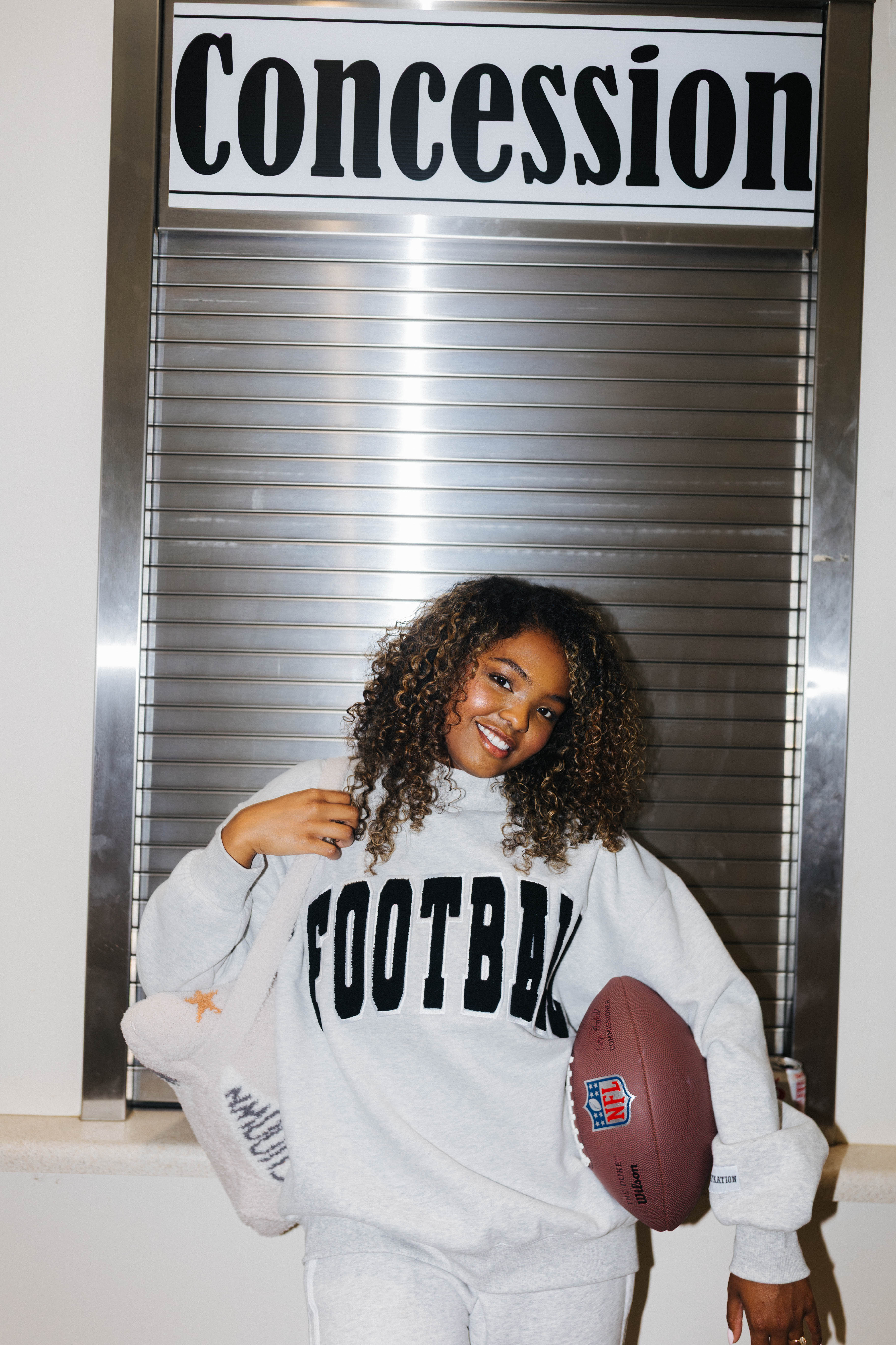 A smiling woman with curly hair stands in front of a metal roll-up door under a sign reading 'Concession', wearing a light gray sweatshirt with 'FOOTBALL' printed in black across the chest and holding a brown football.