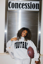 A smiling woman with curly hair stands in front of a metal roll-up door under a sign reading 'Concession', wearing a light gray sweatshirt with 'FOOTBALL' printed in black across the chest and holding a brown football.