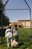 Two young women standing on a grassy baseball field behind a chain-link fence, wearing matching light gray sweatshirts with 'BASEBALL' printed in black on the back. One wears a yellow mit on her head while the other holds the quillow and bat.